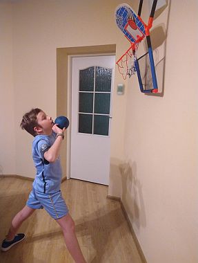 Chłopiec ubrany w strój sportowy rzuca piłkę do kosza zawieszonego na ścianie pokoju.
