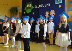 uczniowie klasy 1a w strojach galowych z niebieskimi biretami na głowach stoją w dwuszeregu na tle stosownej dekoracji