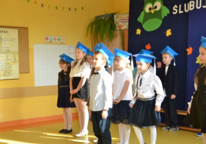 uczniowie klasy 1a w strojach galowych z niebieskimi biretami na głowach stoją w dwuszeregu na tle stosownej dekoracji