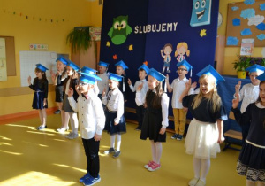 uczniowie klasy 1a w strojach galowych z niebieskimi biretami na głowach stoją w dwuszeregu na tle stosownej dekoracji z uniesioną prawą ręką
