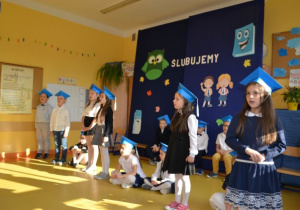 uczniowie klasy 1a w strojach galowych z niebieskimi biretami na głowach na tle stosownej dekoracji