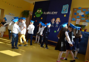 uczniowie klasy 1a w strojach galowych z niebieskimi biretami na głowach tańczą w parach w rozsypce na tle stosownej dekoracji