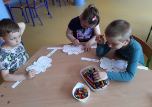 Dzieci siedzą przy stolikach i kolorują opaskę w kształcie motyla.